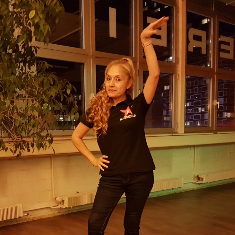 Salsapractica Team Ana Frau in schwarzer Kleidung steht mit hohen SChuhen auf der Tanzfläche und streckt den Arm in die Höhe zu einer Tänzerischen Pose.