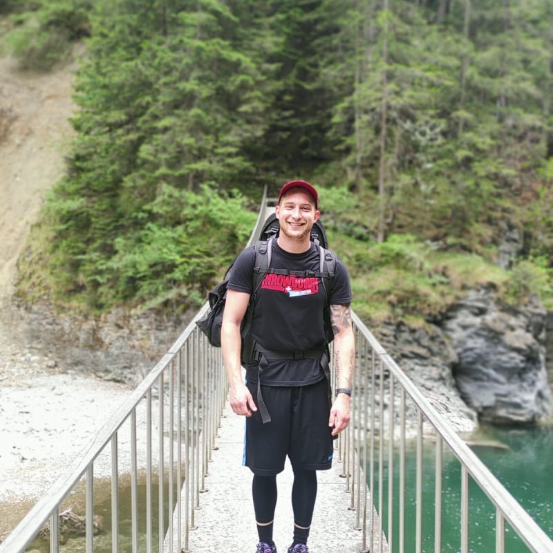 Mann steht auf einer Brücke in Wanderkleidung und einem Hut auf dem Kopf, er lacht und ist fröhlich