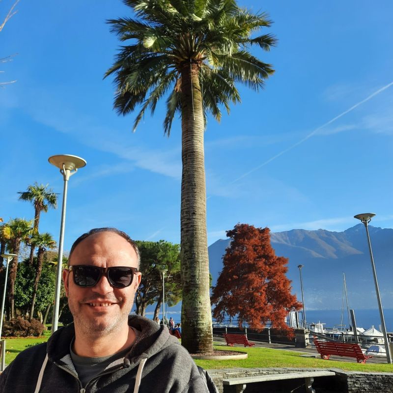 Mann steht mit Sonnenbrille und Sweatjacke im Tessin in der Sonne und lächelt