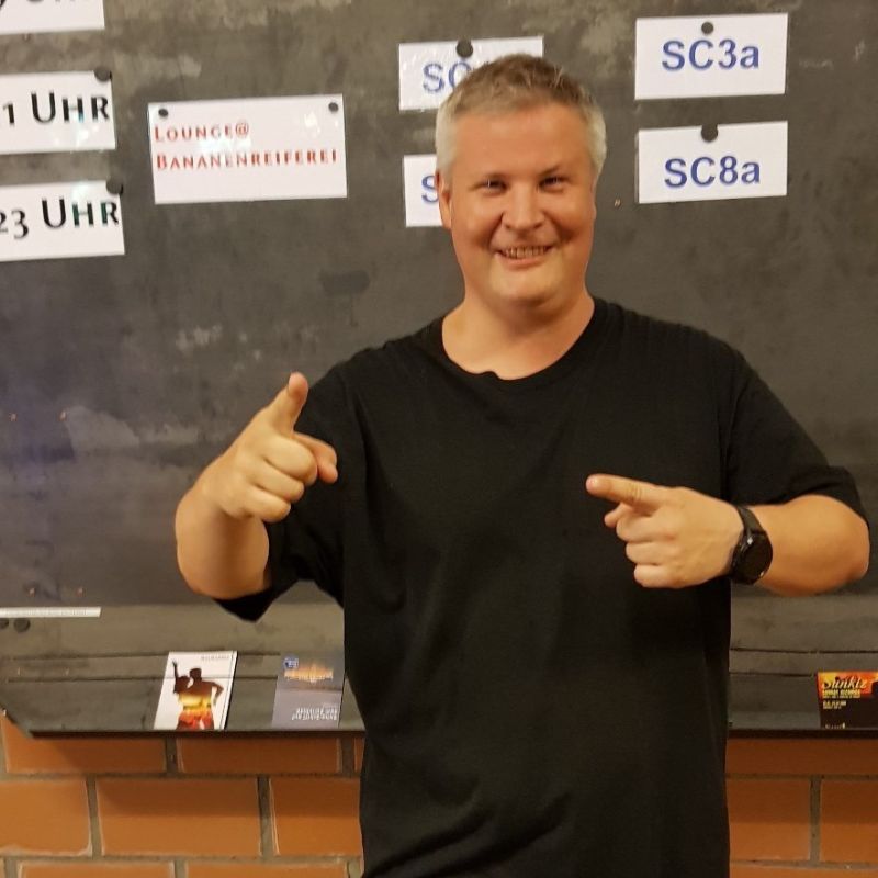 Salsapractica Team Mihael Mann in schwarzem Shirt steht vor einer Wand und zeigt mit den Fingern. Er lacht und hat gute Laune